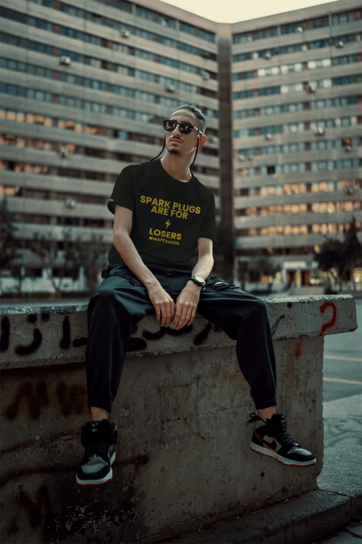Man sitting on a city wall wearing the black Wattz Good "Spark Plugs Are For Losers" EV graphic tee