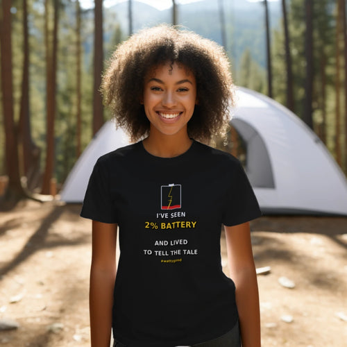 Woman at a campsite wearing the black Wattz Good "I've Seen 2% Battery and Lived to Tell the Tale" EV graphic tee